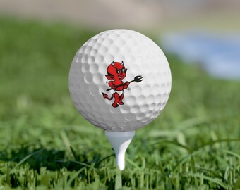 May include: A white golf ball with a red cartoon devil holding a pitchfork printed on it. The golf ball is sitting on a green tee.