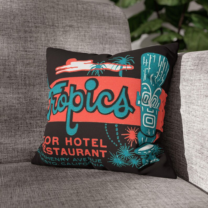 Peut inclure: Coussin d&eacute;coratif noir avec un design r&eacute;tro. Le mot "Tropics" est &eacute;crit en turquoise sur une bande rouge. Un personnage tiki turquoise et des palmiers sont &eacute;galement pr&eacute;sents. Le coussin contient du texte pour un restaurant d'h&ocirc;tel en Californie.