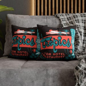 Peut inclure: Deux coussins noirs avec un motif r&eacute;tro "Tropics" en turquoise et corail. Les coussins pr&eacute;sentent un graphisme de palmier et les mots "Motor Hotel Restaurant". Les coussins sont sur un canap&eacute; gris.