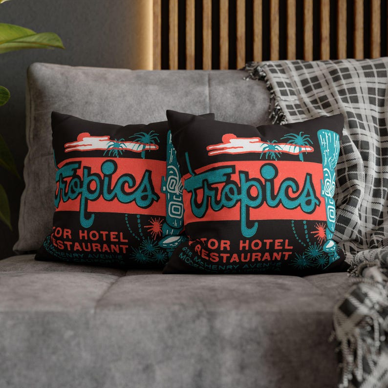Peut inclure: Deux coussins noirs avec un motif r&eacute;tro "Tropics" en turquoise et corail. Les coussins pr&eacute;sentent un graphisme de palmier et les mots "Motor Hotel Restaurant". Les coussins sont sur un canap&eacute; gris.