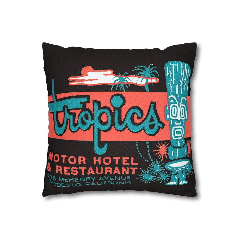 Peut inclure: Coussin carr&eacute; noir avec un design r&eacute;tro. Le coussin pr&eacute;sente le mot "Tropics" en turquoise, sur un rectangle corail. En dessous, il est &eacute;crit "Motor Hotel & Restaurant" avec une adresse. Un personnage tiki turquoise est &agrave; droite.