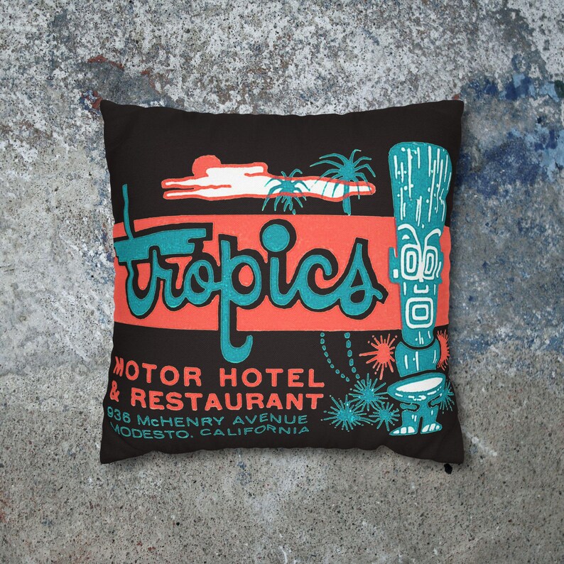 Peut inclure: Coussin carr&eacute; noir avec un design r&eacute;tro. Le mot "Tropics" est en turquoise sur une bande corail. En dessous, le texte indique "Motor Hotel & Restaurant" et l'adresse &agrave; Modesto, Californie. Une figurine tiki turquoise est &agrave; droite.