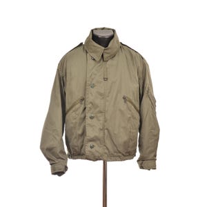 Puede incluir: Chaqueta de estilo militar verde oliva con cuello alto y cierre de botones. La chaqueta tiene dos bolsillos con cremallera en el pecho y dobladillo y puños de canalé. La chaqueta se muestra en un maniquí sobre un fondo blanco.
