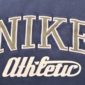 May include: Close-up of a navy blue sweatshirt with the word "NIKE" in large, olive green, arched lettering. Below it, the word "Athletic" is written in a cursive, off-white font. The sweatshirt is a vintage athletic apparel.