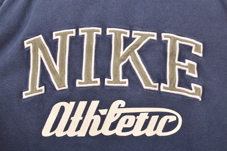May include: Close-up of a navy blue sweatshirt with the word "NIKE" in large, olive green, arched lettering. Below it, the word "Athletic" is written in a cursive, off-white font. The sweatshirt is a vintage athletic apparel.