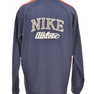 May include: Navy blue long-sleeve shirt with the Nike logo and "Athletic" text on the back. The shirt has a cream-colored collar and red accents on the shoulders. The shirt is displayed on a mannequin.