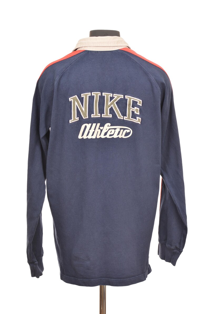 May include: Navy blue long-sleeve shirt with the Nike logo and "Athletic" text on the back. The shirt has a cream-colored collar and red accents on the shoulders. The shirt is displayed on a mannequin.