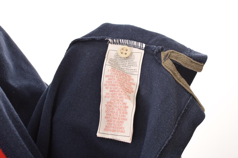 May include: Navy blue polo shirt with a red stripe detail. The shirt features a button-down collar and a tag with text. The collar has a brown trim.