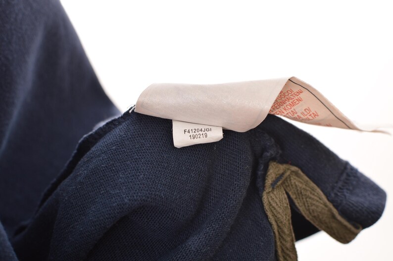 May include: Close-up of a navy blue garment with a tag attached. The tag has the text "F41204JGI 190219" printed on it. The garment has a textured appearance and a contrasting olive green trim.