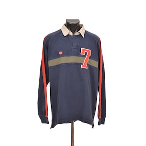 May include: Navy blue long-sleeve rugby shirt with a white collar and red accents on the sleeves. Features a green stripe across the chest and a large red number 7. The shirt has a classic, sporty design.
