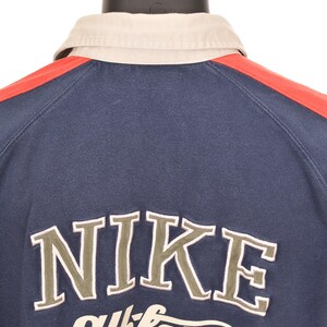 May include: Navy blue athletic shirt with a beige collar and red shoulder stripes. The word "NIKE" is embroidered in a light green color, with "athletic" underneath. The shirt is displayed on a black mannequin.