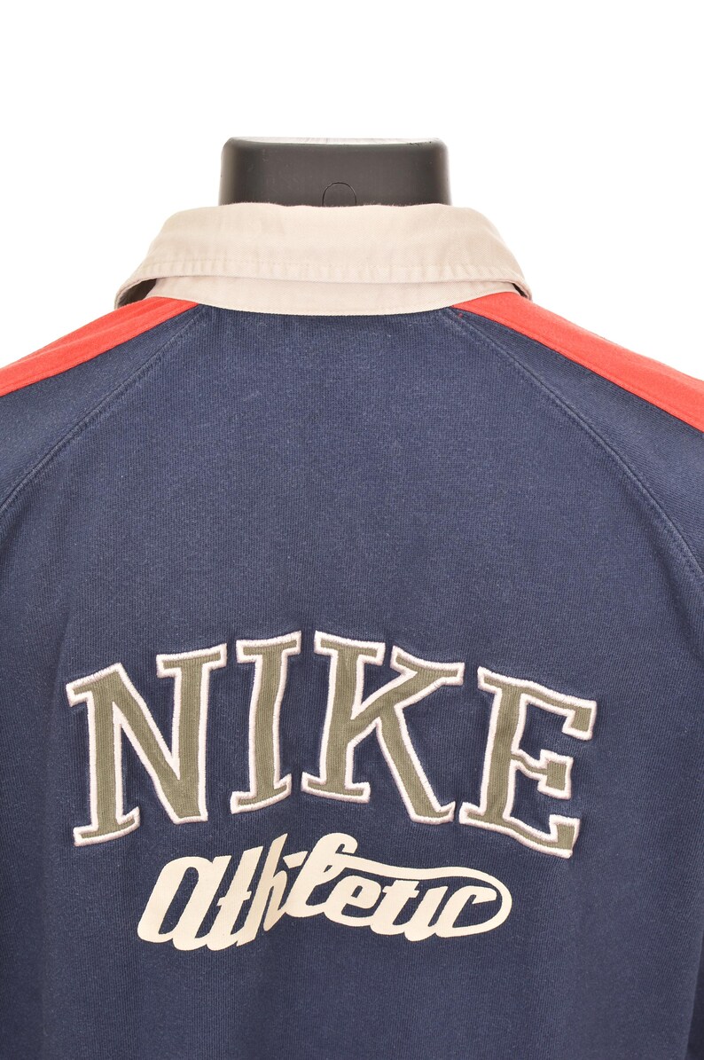May include: Navy blue athletic shirt with a beige collar and red shoulder stripes. The word "NIKE" is embroidered in a light green color, with "athletic" underneath. The shirt is displayed on a black mannequin.