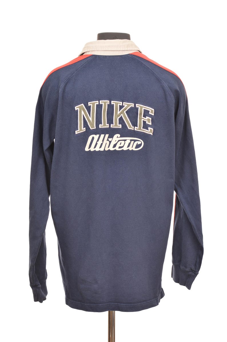 May include: Navy blue long-sleeve shirt with the Nike logo and "Athletic" in white lettering on the back. The shirt has a beige collar and red accents on the shoulders. A vintage athletic top.
