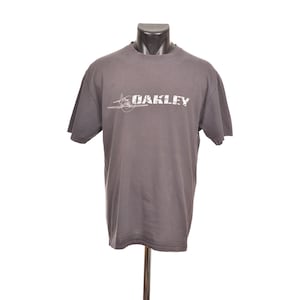 May include: Gray short-sleeve t-shirt with the white text "OAKLEY" and a graphic of a propeller plane. The shirt is displayed on a mannequin.