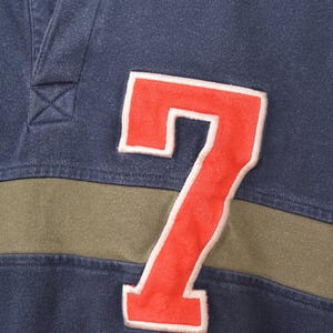 May include: Close-up of a navy blue rugby shirt with a cream-colored collar and a red number 7 patch. The shirt has an olive green stripe across the chest. The shirt is made of a soft, comfortable material.