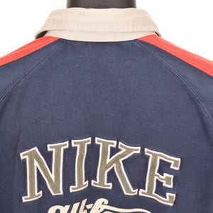 May include: Navy blue athletic shirt with a beige collar and red shoulder stripes. The word "NIKE" is embroidered in a light green color, with "athletic" underneath. The shirt is displayed on a black mannequin.