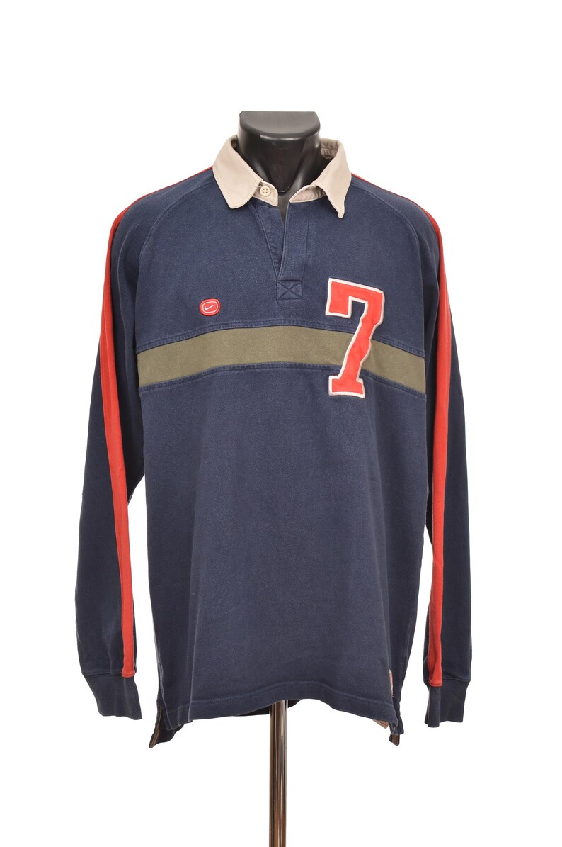 May include: Navy blue long-sleeve rugby shirt with a beige collar and red stripes down the sleeves. Features a green horizontal stripe across the chest and a large white outlined red number 7. The shirt has a button-up collar.