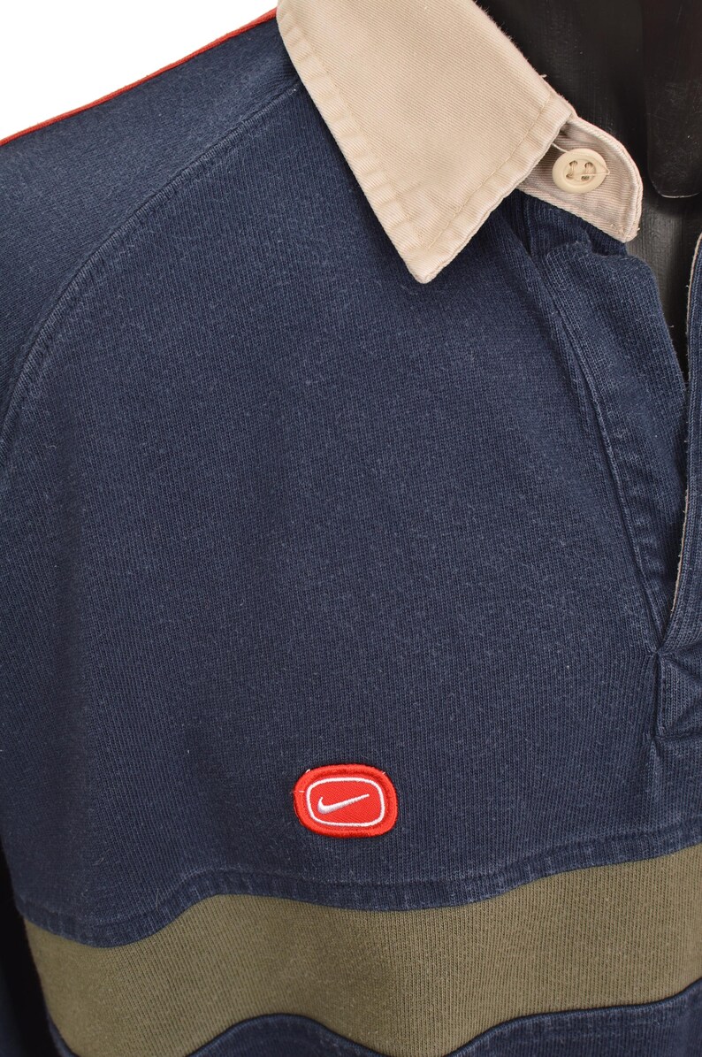 May include: Navy blue long-sleeve rugby shirt with a tan collar and red trim. Features a green horizontal stripe across the chest and a red Nike logo. The shirt has a buttoned placket.