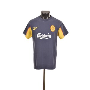May include: A dark navy blue short-sleeve t-shirt featuring a white Carlsberg logo across the chest. The shirt includes a yellow Reebok logo on the right side and a circular red and yellow emblem on the left side. The sleeves have yellow panels.
