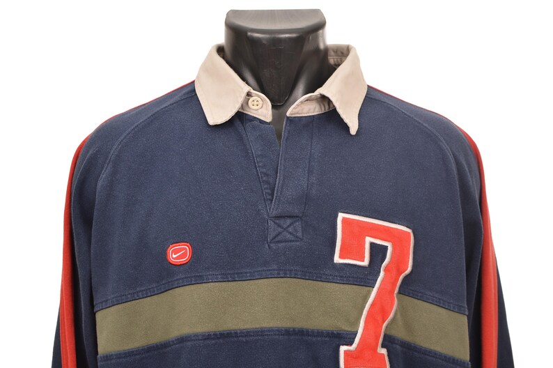 May include: Navy blue long-sleeve shirt with a beige collar and red sleeves. Features a red "7" on the front and a small red Nike logo. The shirt has a green stripe across the chest.