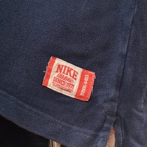 May include: Close-up of a navy blue t-shirt with a red and white Nike label. The label reads "NIKE Athletic SINCE 1971" and includes the address "ONE BOWERMAN DRIVE BEAVERTON OREGON".