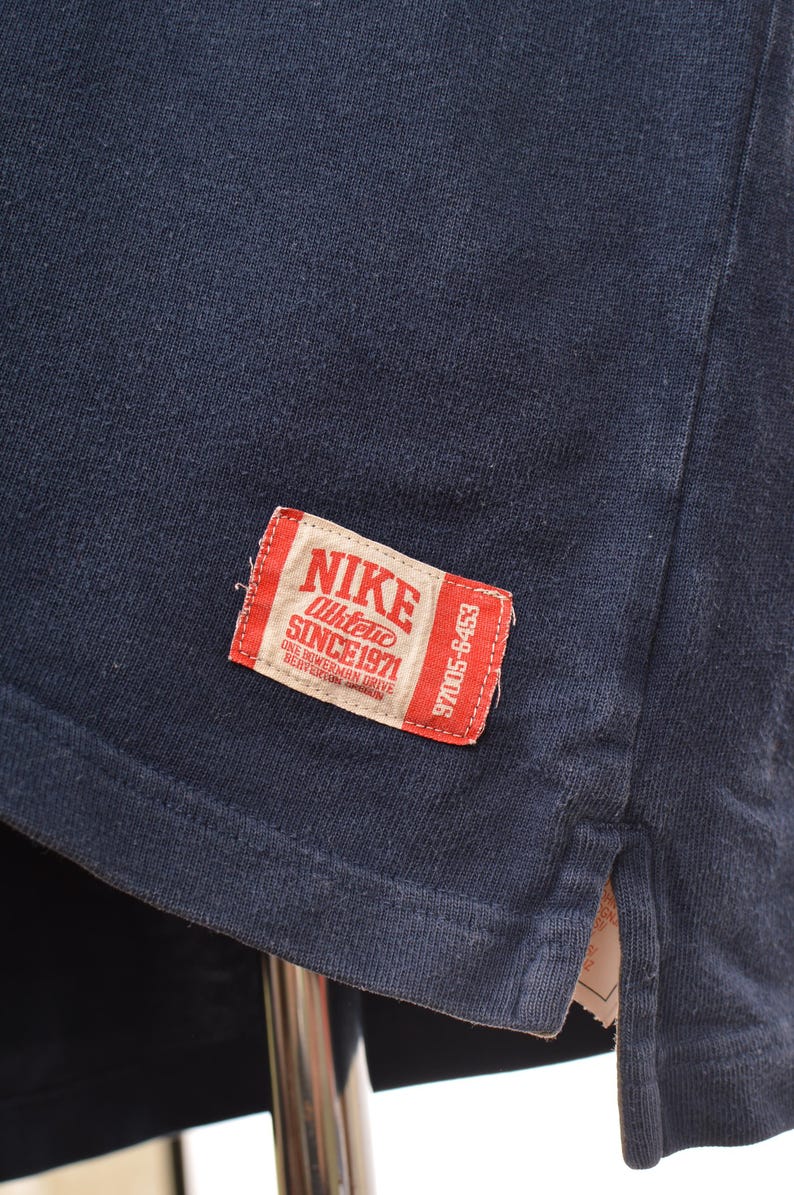 May include: Close-up of a navy blue t-shirt with a red and white Nike label. The label reads "NIKE Athletic SINCE 1971" and includes the address "ONE BOWERMAN DRIVE BEAVERTON OREGON".
