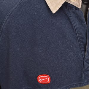 May include: Navy blue long-sleeve rugby shirt with a tan collar and red trim. Features a green horizontal stripe across the chest and a red Nike logo. The shirt has a buttoned placket.