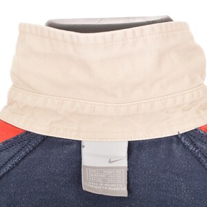 May include: Close-up of a navy blue and red athletic top with a cream-colored collar. The top features a tag with the Nike logo and size information. The collar is folded over and the top is made of a soft material.