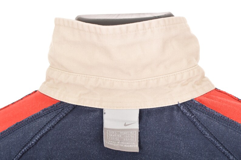 May include: Close-up of a navy blue and red athletic top with a cream-colored collar. The top features a tag with the Nike logo and size information. The collar is folded over and the top is made of a soft material.