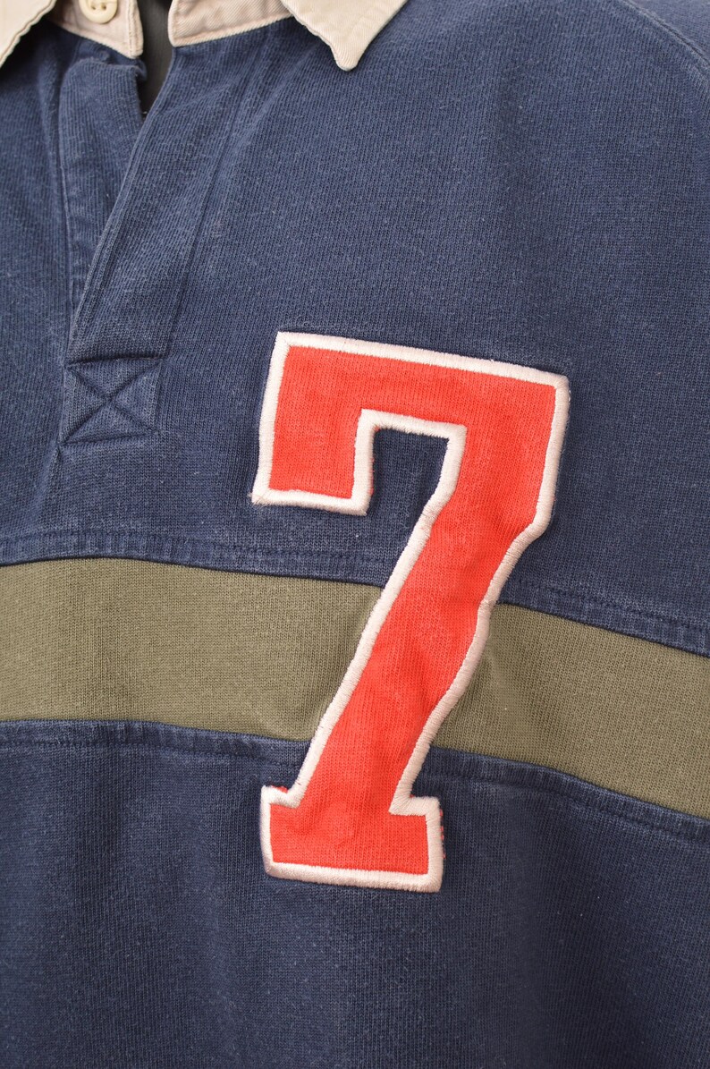 May include: Close-up of a navy blue rugby shirt with a cream-colored collar and a red number 7 patch. The shirt has an olive green stripe across the chest. The shirt is made of a soft, comfortable material.