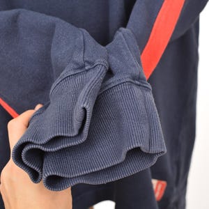 May include: Close-up of a navy blue sweatshirt sleeve with a ribbed cuff. A bright red stripe runs down the sleeve. The fabric appears to be a soft, comfortable material.