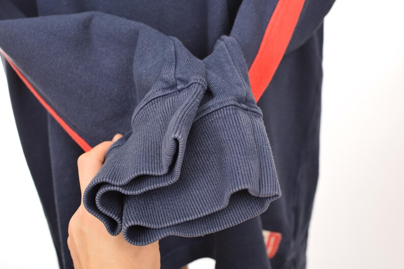 May include: Close-up of a navy blue sweatshirt sleeve with a ribbed cuff. A bright red stripe runs down the sleeve. The fabric appears to be a soft, comfortable material.