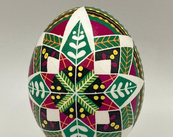 May include: A decorative egg with a geometric pattern in white, green, magenta, and yellow, displayed on a black stand. The egg features a central starburst design and leaf motifs, creating a vibrant and detailed appearance.