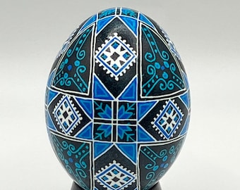 May include: An intricately decorated egg featuring a geometric pattern in blue, white, and black. The egg is displayed on a black stand. The design includes diamond and star shapes. The egg's patterns are detailed and symmetrical.
