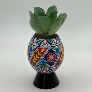 Puede incluir: Una maceta en forma de huevo con un diseño de arte popular colorido, con patrones rojos, azules y amarillos. Contiene una planta suculenta verde. La maceta se apoya en una base negra.
