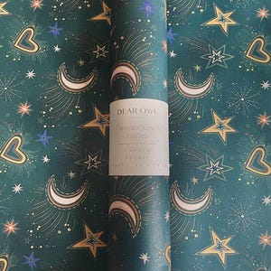May include: A roll of dark teal wrapping paper with a celestial pattern. The design features gold stars, crescent moons, and hearts, along with blue and white accents. The paper has a label that reads "DEAR OWL" in English.