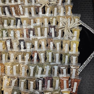 May include: An assortment of small, clear plastic bags filled with various dried herbs and spices. Each bag has a white label with text. Two bundles of dried sage are placed on top of the bags. The background is a black surface.