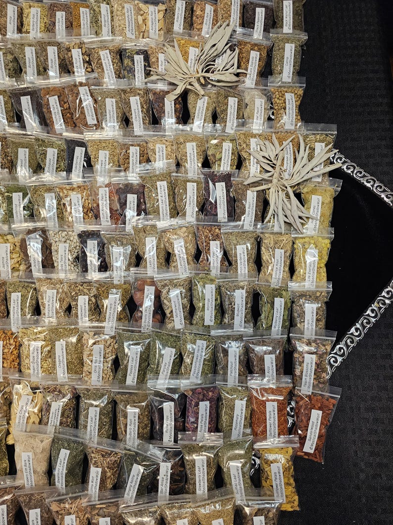 May include: An assortment of small, clear plastic bags filled with various dried herbs and spices. Each bag has a white label with text. Two bundles of dried sage are placed on top of the bags. The background is a black surface.