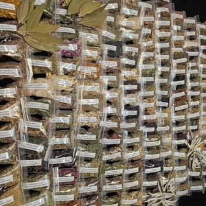 May include: A display of various dried herbs and spices in small, clear plastic bags. Large, dried bay leaves are arranged on top. Each bag has a label with the herb's name. The arrangement is on a black surface, showcasing a wide variety of culinary and medicinal ingredients.