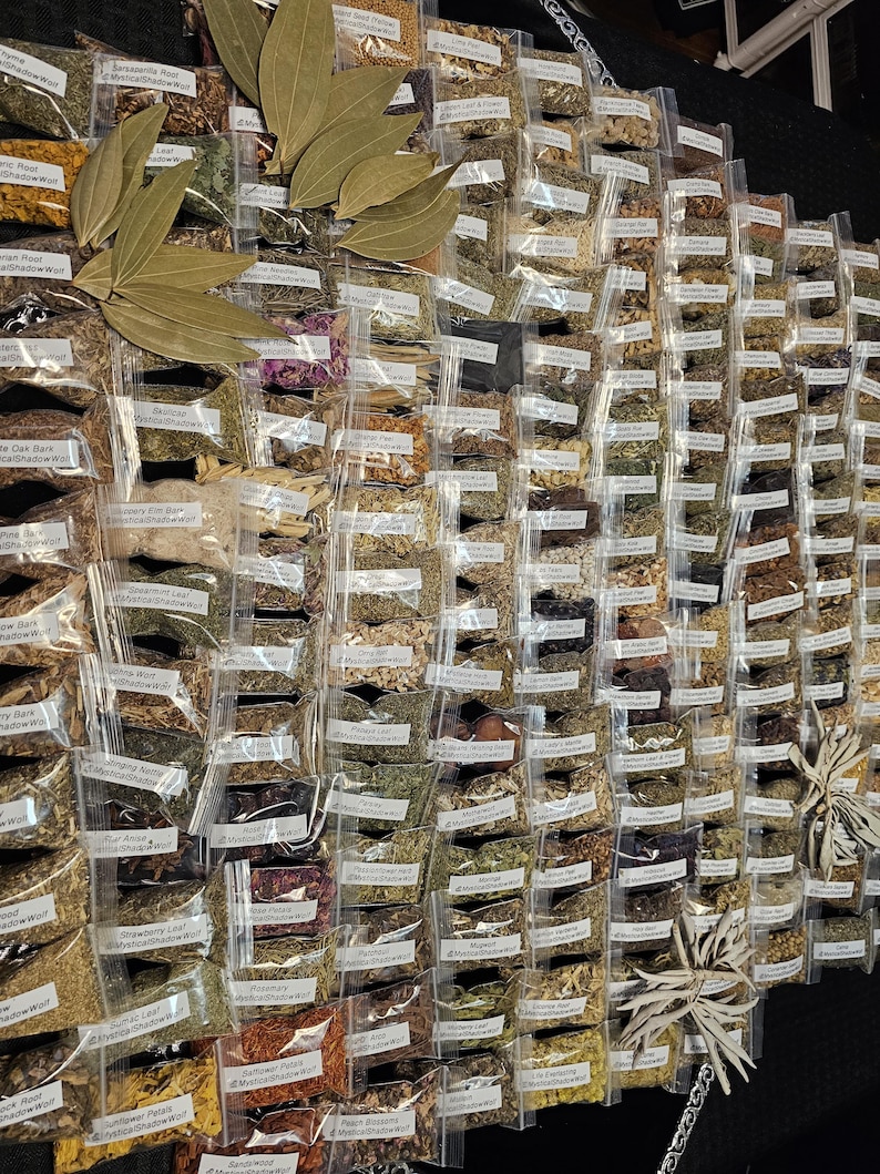 May include: A display of various dried herbs and spices in small, clear plastic bags. Large, dried bay leaves are arranged on top. Each bag has a label with the herb's name. The arrangement is on a black surface, showcasing a wide variety of culinary and medicinal ingredients.