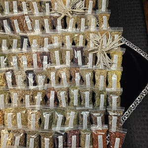 May include: An assortment of small, clear plastic bags filled with various dried herbs and spices. Each bag has a white label with black text. The bags are arranged in a grid pattern, with a few decorative dried plants interspersed throughout.