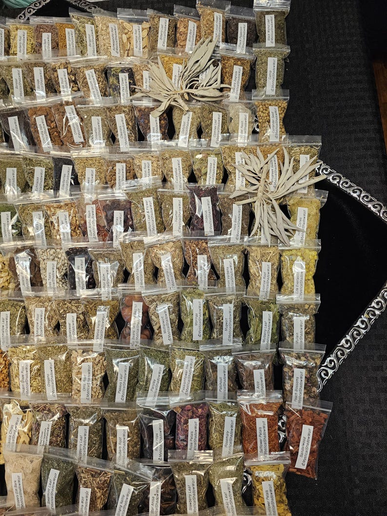 May include: An assortment of small, clear plastic bags filled with various dried herbs and spices. Each bag has a white label with black text. The bags are arranged in a grid pattern, with a few decorative dried plants interspersed throughout.