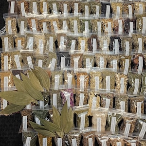 May include: An overhead shot of numerous small, clear plastic bags filled with various dried herbs and spices. Several large, dried bay leaves are arranged in the foreground. Each bag has a small label with text. The collection is arranged on a dark surface.