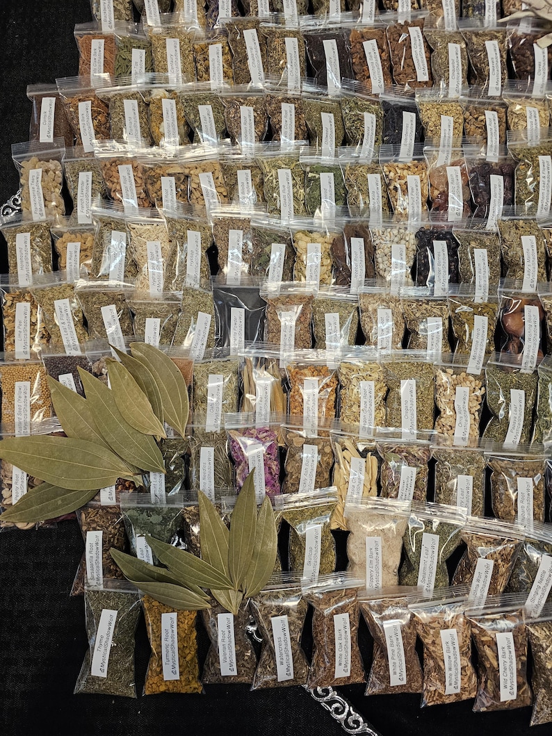 May include: An overhead shot of numerous small, clear plastic bags filled with various dried herbs and spices. Several large, dried bay leaves are arranged in the foreground. Each bag has a small label with text. The collection is arranged on a dark surface.
