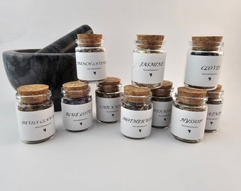 May include: A collection of small glass jars with cork lids filled with dried herbs and flowers. The jars are labeled with the names of the herbs and flowers, including French Lavender, Jasmine, Cloves, Devil's Claw, Blue Lotus, Chickweed, Motherwort, Peppermint, Hyssop, and  Rosemary.
