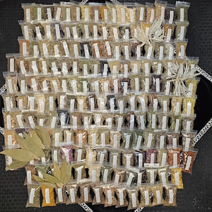 May include: A collection of small, clear plastic bags filled with various dried herbs and spices, arranged in a grid pattern. The bags have white labels. Several bundles of dried leaves are placed around the bags. The herbs and spices are in shades of brown, green, and yellow.