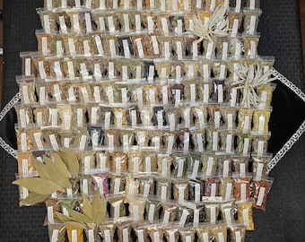 May include: A collection of small, clear plastic bags filled with various dried herbs and spices, arranged in a grid pattern. The bags have white labels. Several bundles of dried leaves are placed around the bags. The herbs and spices are in shades of brown, green, and yellow.