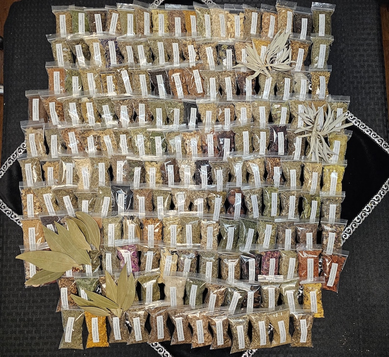 May include: A collection of small, clear plastic bags filled with various dried herbs and spices, arranged in a grid pattern. The bags have white labels. Several bundles of dried leaves are placed around the bags. The herbs and spices are in shades of brown, green, and yellow.