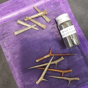 May include: A collection of items on a purple drawstring bag. Includes metal nails, a small jar labeled "Black Salt Mystical Shadow", and several thorny branches. The items are arranged on a dark surface.