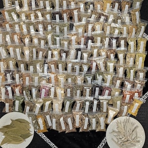 May include: A large collection of small, clear plastic bags filled with various dried herbs and spices. The bags are arranged in a dense pattern, with two white plates holding bay leaves and sage at the bottom. The background is a black surface.